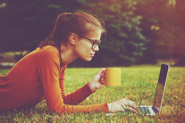 girl lying on the grass and using laptop and typing with coffee