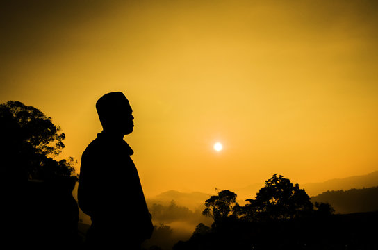 silhouette image of a man standing on top off the hill. sunset sunrise. dark tree as background