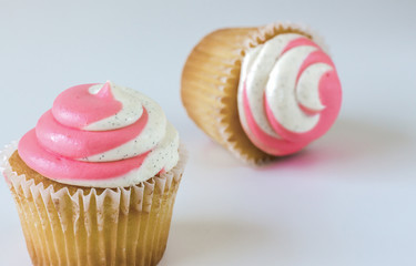 horizontal image of two white cupcakes with pink and white swirl icing with one standing upright and the other lying on its side on white background