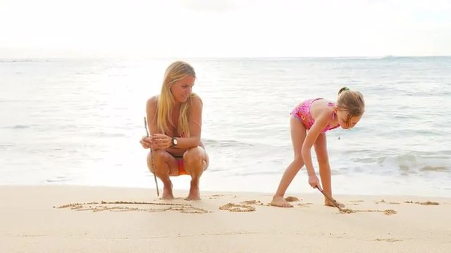 Blonde mother and daughter writing in sand on a beach