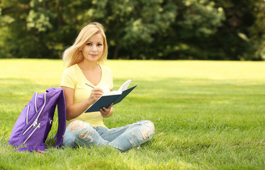 Naklejka na ściany i meble Student girl with book and backpack in park. Blonde young woman.