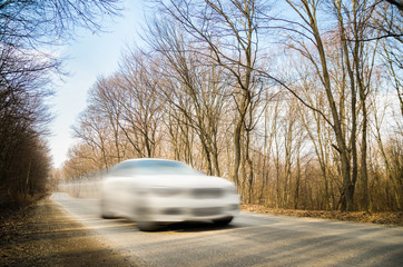 Speeding car with a motion look on a road through a leafless forest on a sunny autumn day. Soft filters applied