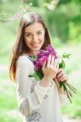 Happy lifestyle concept with attractive smiling young woman holding a bunch of spring blooming lilac close to her face