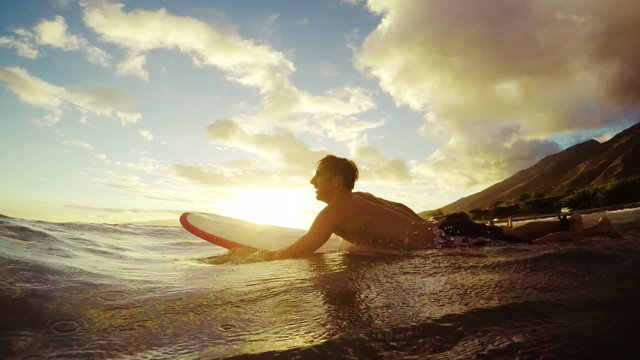 Young Man Paddles a Surfboard Through a Golden Sunset. View From in the Water.