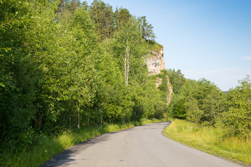 Fototapeta na wymiar Tourist route to the Urals. Asphalt road leads to the rock in the green forest on background blue sky 