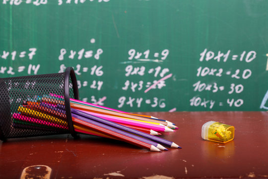 Colorful pencils of red yellow orange violet purple pink green and blue in stationary cup and steel lying on brown school desk on written with white chalk blackboard backgroung on lesson of math