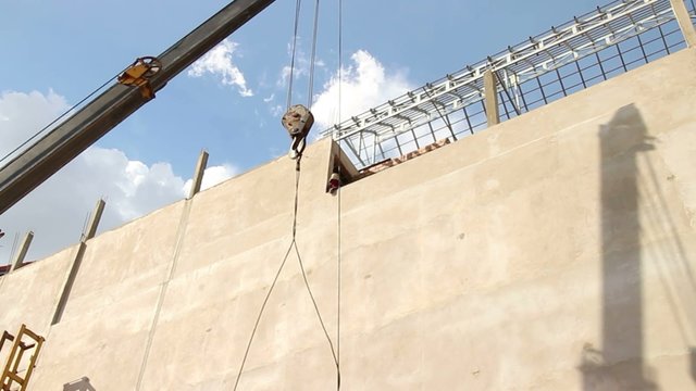 wide shot of a long concrete pile being lifted by a crane from a resting horizontal position up to a vertical position; hooks and pulley dangle, with strong shadows against opposite building.