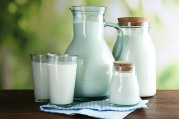 Pitcher, jars and glasses of milk on wooden table, on nature background