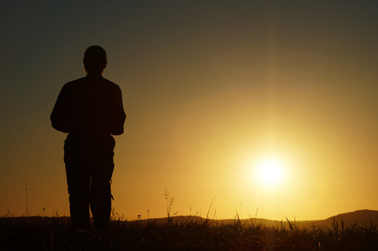 Silhouette of a man on a grassy horizon at sunset. Wooded mountains in the background.