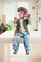Cute little boy in vintage outfit sitting on a table and eating strawberry while playing with a tin can telephone