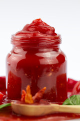 Tomato ketchup in a glass jar and green leaves on white . Homemade, healthy vegetarian food
