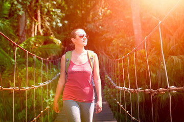 Happy smiling woman hiker crossing suspension bridge in sunlight - obrazy, fototapety, plakaty