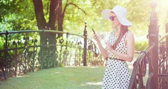 Woman using smartphone in the park. Beautiful smiling attractive lady wearing white hat and using modern phone in sunny green outdoors. Communication and technology concept. 4K, DCi, slow motion.