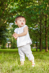 Portrait of cute adorable little Asian girl child baby one year old in white pants shirt, standing on field meadow grass on sunset playing clapping her hands