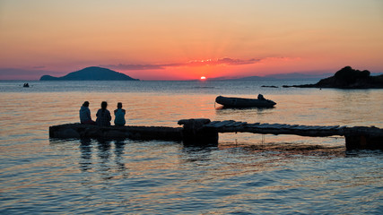 Girls sitting on a pier in front of small island at sunset