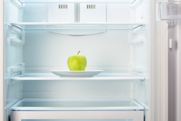 Green apple on white plate in open empty refrigerator