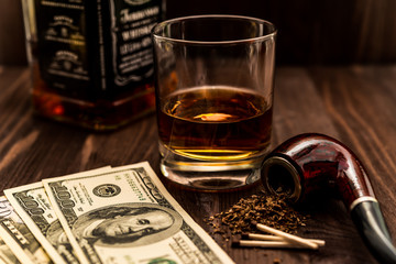 Glass of whiskey with a money and tobacco pipe with tobacco leaves on a wooden table