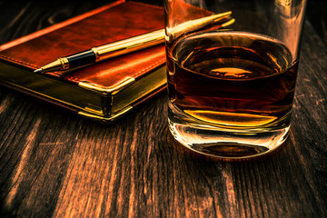 Glass of whiskey and  leather notebook with golden pen on a wooden table