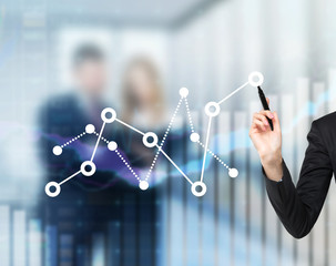A woman's hand is drawing fluctuation of the financial chart. A couple of colleagues on the background in blur.
