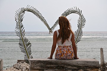 A young single woman is sitting on the beach in front of a big heart without a relationship or a partner / boyfriend. This makes her sad, because she is alone / lonely and searches for love.