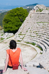 Tourist girl with an orange t-shirt sitting on one of the steps of the Segesta theather, West Sicily, and looking at the view