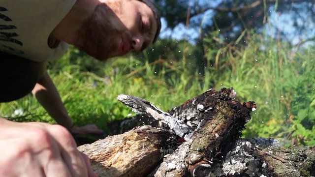 Man fanning the flames of a fire in nature. Video with sound. You can hear the crackle of twigs, sound fanning the flames, the birds singing, the sound of the river.
