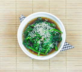 Homemade stir fried spinach with coconut butter, garlic, soy sauce and sesame seeds in a white bowl on wooden background.