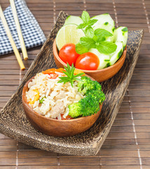 Healthy eating, homemade stir fried rice with vegetable, meat and egg served on wooden bowl and side bowl of fresh cucumbers, tomatoes on wooden plate with chopsticks (Selective Focus, Focus on rice).