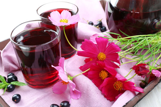 Glasses of fresh blackcurrant juice on wooden tray with pink napkin and flowers, closeup