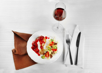 Dish of baked chicken leg and vegetable salad in white plate with glass of wine on table with napkin, closeup