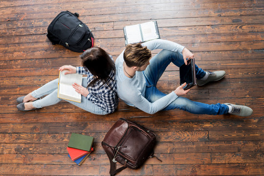 Girl reading a book and boy using a tablet leaning on each other on wooden floor having notebooks and bags around them.