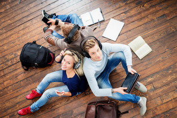 Group of students using smartphones and tablet in headphones listening to the music and leaning on each other on wooden floor having notebooks and bags around them.
