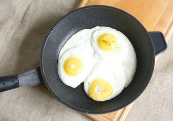 Three fried eggs in a pan on a wooden board and gray sackcloth