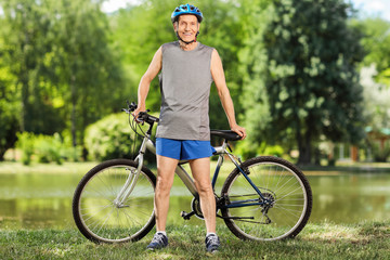 Naklejka premium Cheerful senior cyclist posing in front of his bicycle by a pond