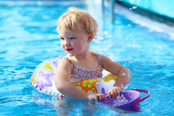 Adorable happy little child, curly toddler girl in swimming suit having fun relaxing and floating on an inflatable toy ring in a pool on sunny day during summer vacation in resort