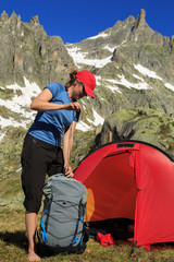 Hiker woman packing her backpack in front of a red tent in the Swiss mountains. Outdoors summer trekking vacation concept.