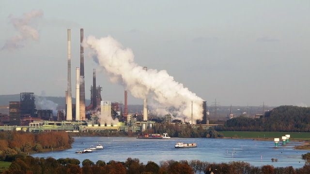 A large steaming coking plant in the distance with some ships on Rhine River in the foreground. Shot in Moers with view to Duisburg, Germany.