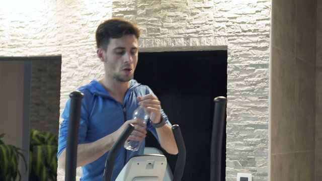 Young man exercising on elliptical machine in the gym and drinking water 
