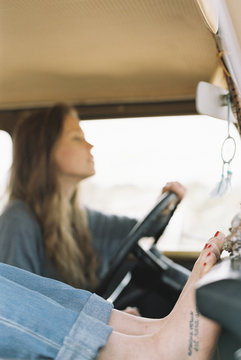 Barefoot woman resting her feet on the dashboard of a 4x4, a tattoo on her right foot, another woman driving.