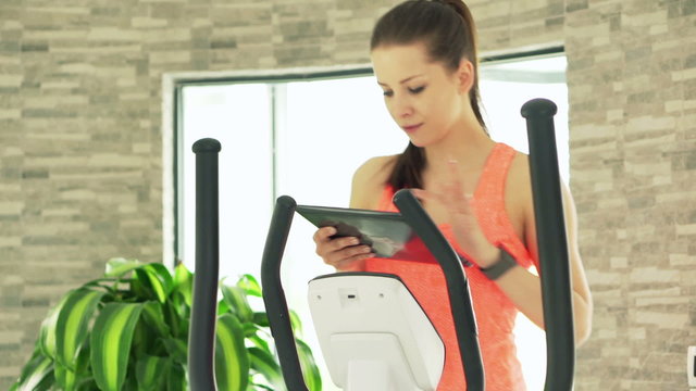 Young woman with tablet computer exercising on elliptical machine in gym

