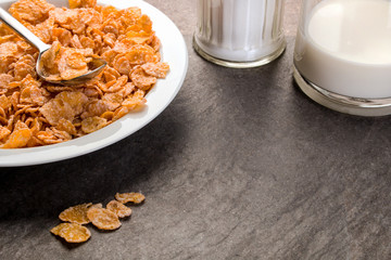 Corn Flakes auf einem Teller, Milch in einer Flasche und ein Zuckerstreuer auf einer schwarzen Granitplatte