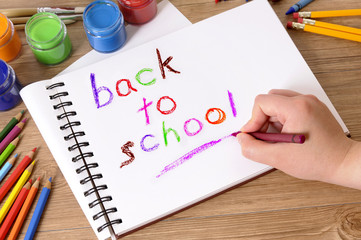 Child hand writing back to school word with crayons on open notebook book student desk surrounded by pencils photo