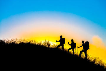 Fototapeta na wymiar Family journey wild landscape Silhouettes of three people walking with backpacks other hiking gear up toward top of wild grass mountain mother father daughter bright luminous sunrise sky background