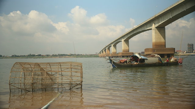 Small wooden fishing boat on the river nearby a bridge; Traditional bamboo fish trap in the foreground