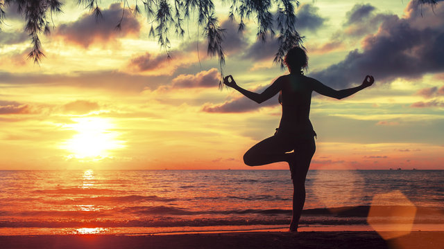 Silhouette of young woman practicing yoga on the beach at amazing sunset.
