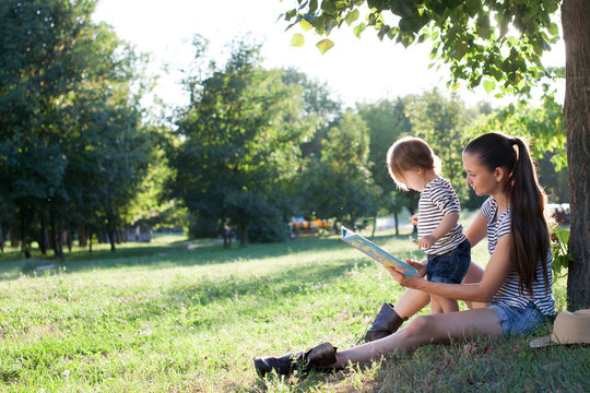 Young Stylish Mother And Toddler Reading Book At Garden During Summer Fun