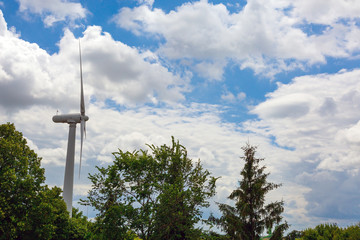 Wind energy.
Image of single white wind turbine located among forest tree with massive clouds and blue sky background