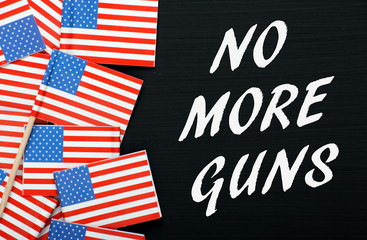 The phrase No More Guns in white text on a blackboard next to flags of the United States of America as a call for gun control