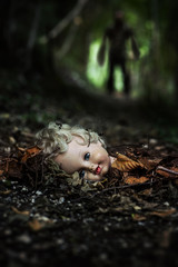 A doll's head lying in the middle of a path of a dark forest. There is a haunting presence in the background blurred.
