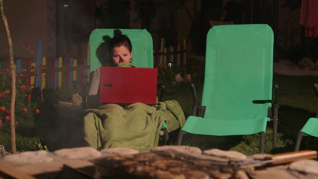 Young girl wrapped by rug sitting on lounger in backyard with laptop
